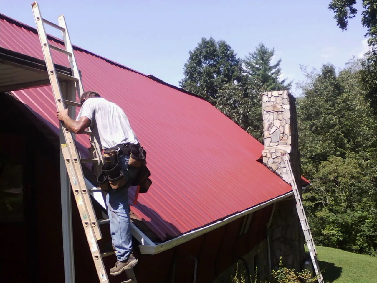 Roofer on a ladder beside a red metal roof and stone chimney — Soffit & Fascia Repair by Roofing Brothers in Boring